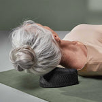 Eine ältere Frau mit grauem Haar in einem Dutt liegt auf dem Rücken auf einer Matte und benutzt ein schwarzes Nackenstützkissen - perfekt, um "Nackenschmerzen selbst behandeln" von Liebscher & Bracht zu üben, um Nackenschmerzen zu lindern.