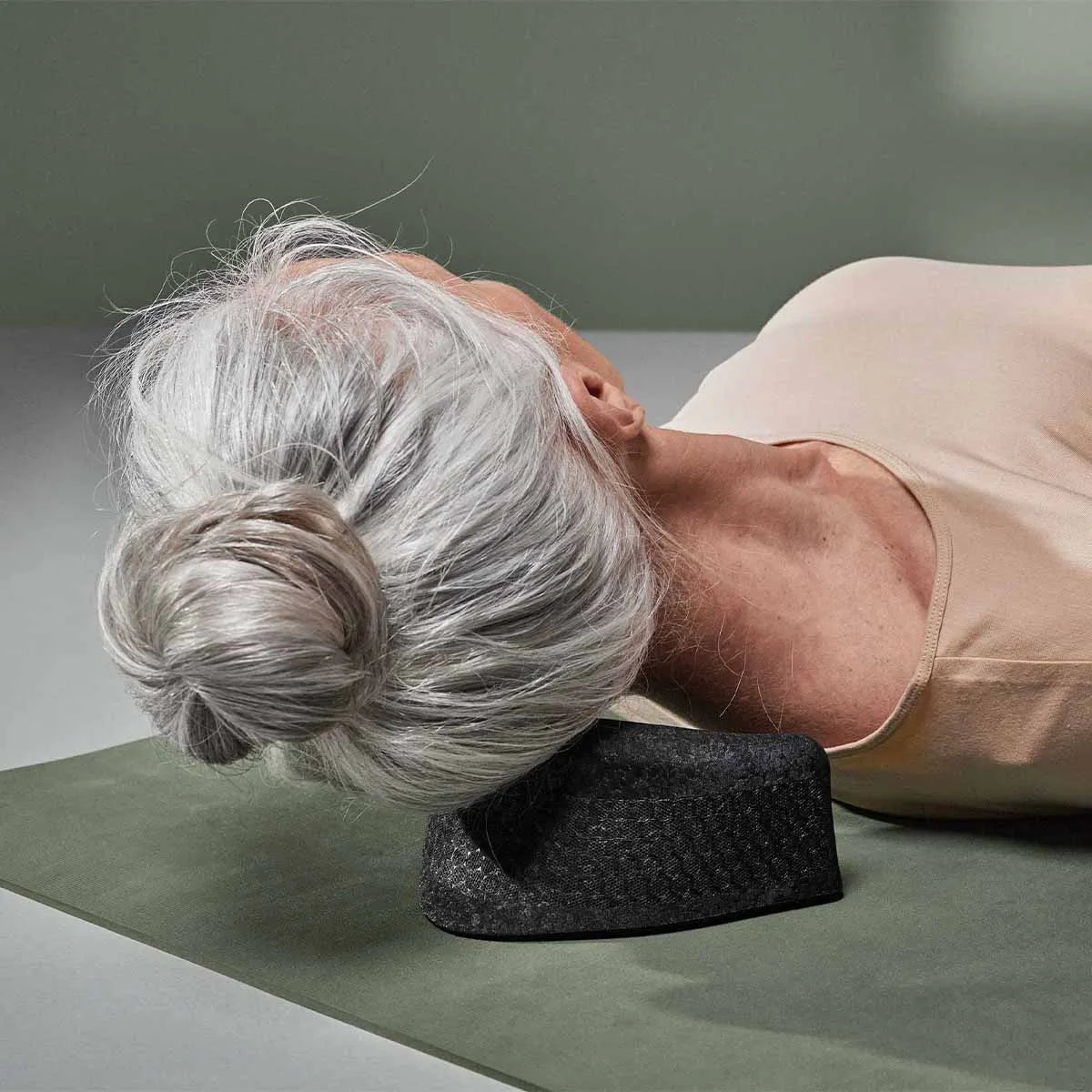 Eine ältere Frau mit grauem Haar in einem Dutt liegt auf dem Rücken auf einer Matte und benutzt ein schwarzes Nackenstützkissen - perfekt, um "Nackenschmerzen selbst behandeln" von Liebscher & Bracht zu üben, um Nackenschmerzen zu lindern.