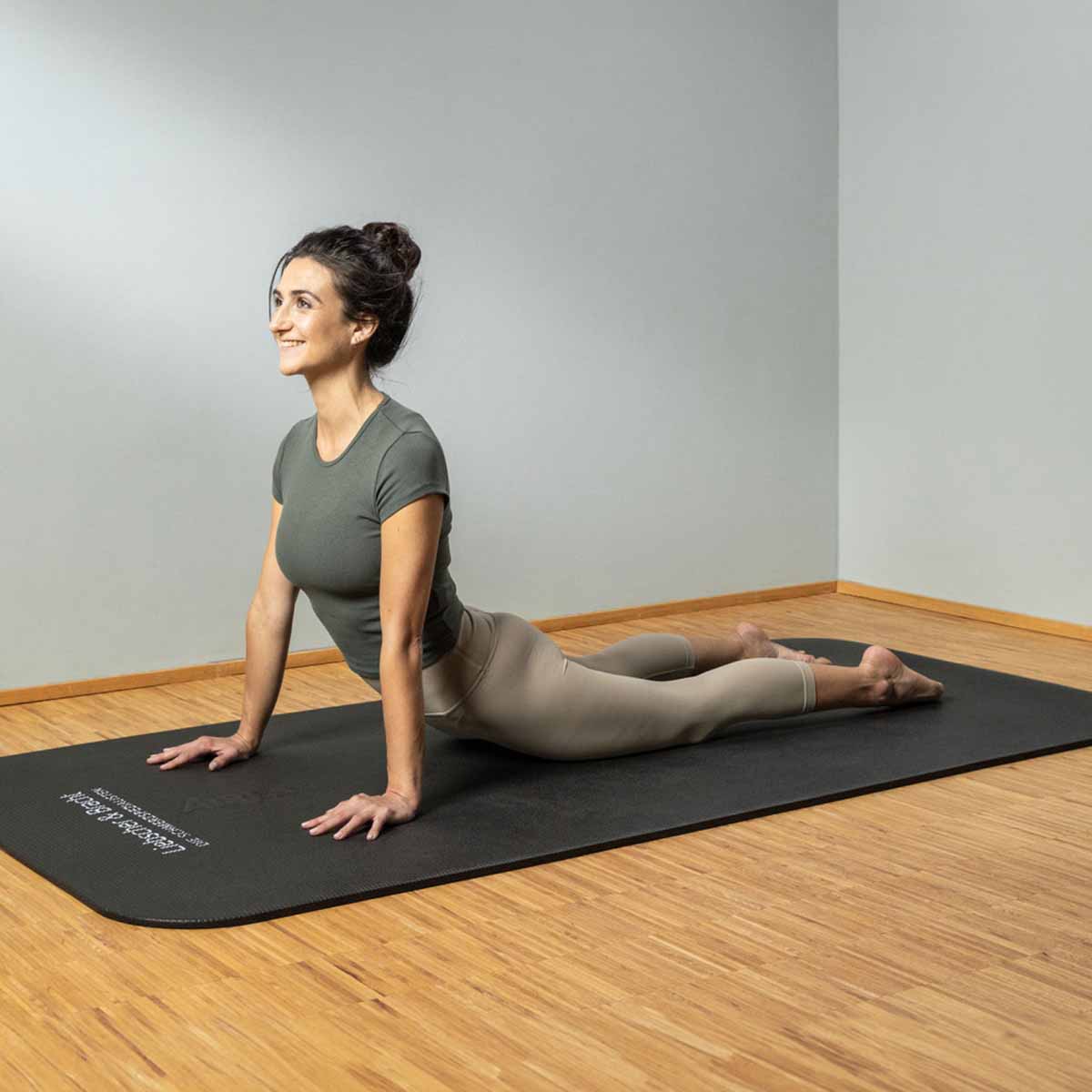 Eine Frau übt drinnen auf einer schwarzen Matte Yoga und nimmt die Kobra-Pose ein, wobei sie die Arme gerade hält, die Brust hebt und die Beine hinter sich ausstreckt, während sie lächelt.