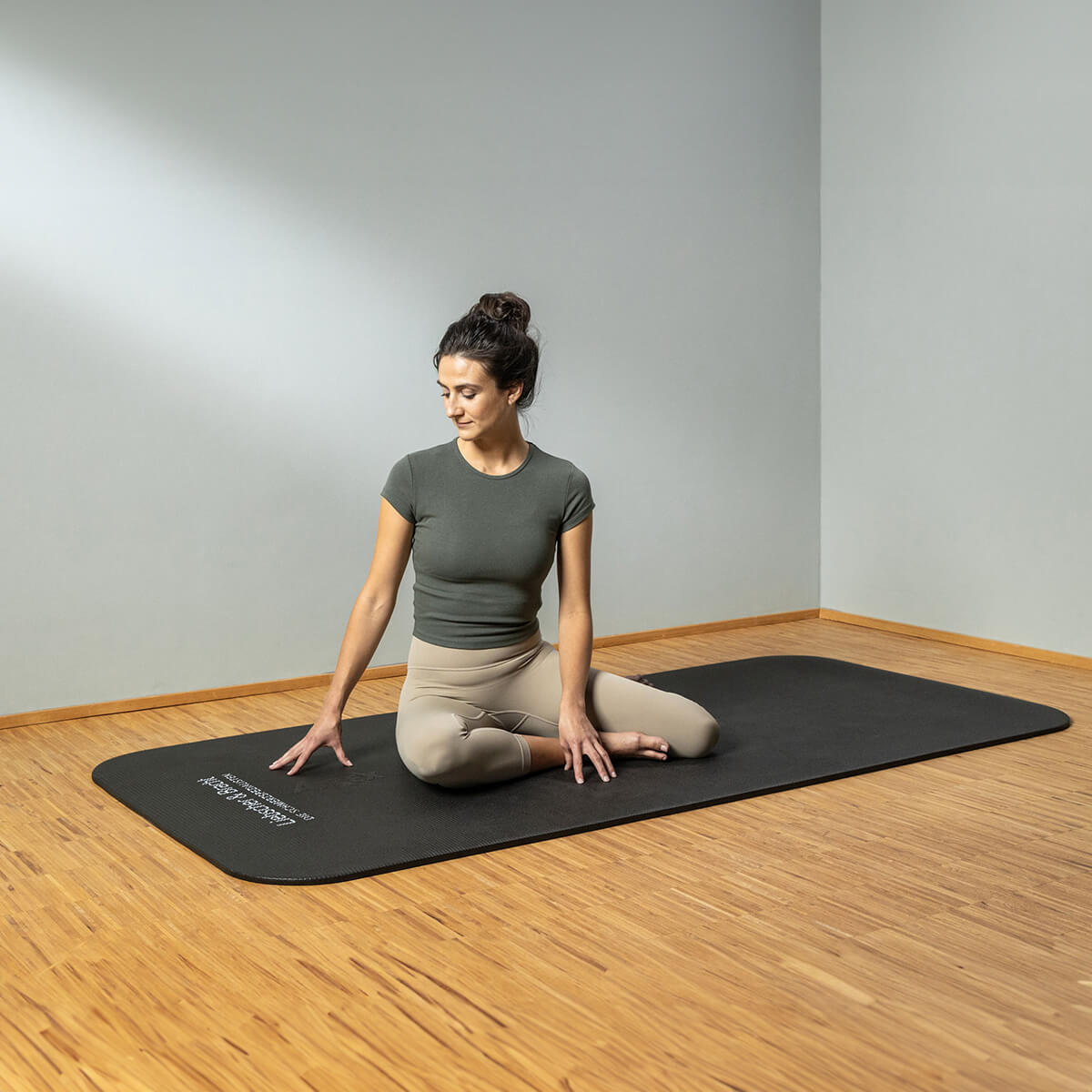 In einem hellen, minimalistischen Raum mit Holzfußboden und hellen Wänden sitzt eine Frau auf einer schwarzen Yogamatte in einer knienden Drehung, eine Hand auf der Matte hinter sich und die andere auf ihrem Knie.