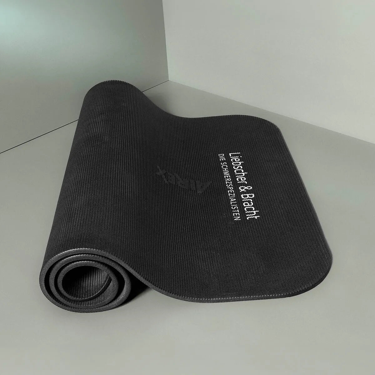 Eine schwarze Rolands Übungsmatte von Liebscher & Bracht wird auf einer grauen, rutschfesten Unterlage teilweise abgerollt. Auf der Matte sind "Leichter bei Bracht" und ein kleinerer weißer Text aufgedruckt.