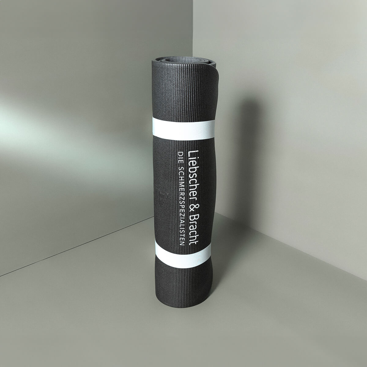 Eine aufgerollte schwarze Gymnastikmatte mit weißer Umrandung, auf der in weißer Schrift der Text Liebscher & Bracht und DIE SCHMERZSPEZIALISTEN zu lesen ist, steht aufrecht in einer Ecke auf einer grauen Fläche.