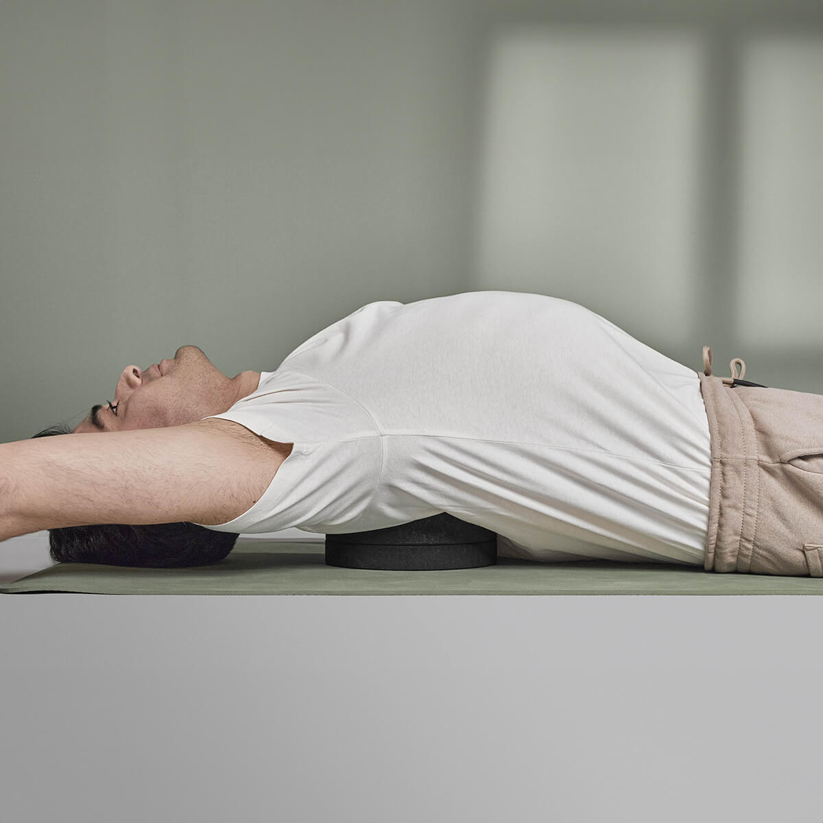 Die Person liegt auf dem Rücken, die Arme sind über Kopf ausgestreckt. Der Rückenretter von Liebscher & Bracht wird unter dem oberen Rücken eingesetzt, um die Entspannung und sanfte Mobilisierung der Rückenmuskulatur zu fördern.