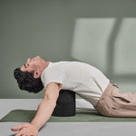 Ein Mann in weißem T-Shirt und beiger Hose benutzt ein Yoga-Bolster von Liebscher & Bracht, während er Übungen aus "Rücken- & Bandscheibenschmerzen selbst behandeln" auf einer grünen Matte in einem sonnenbeschienenen Raum mit grünen Wänden vorträgt.