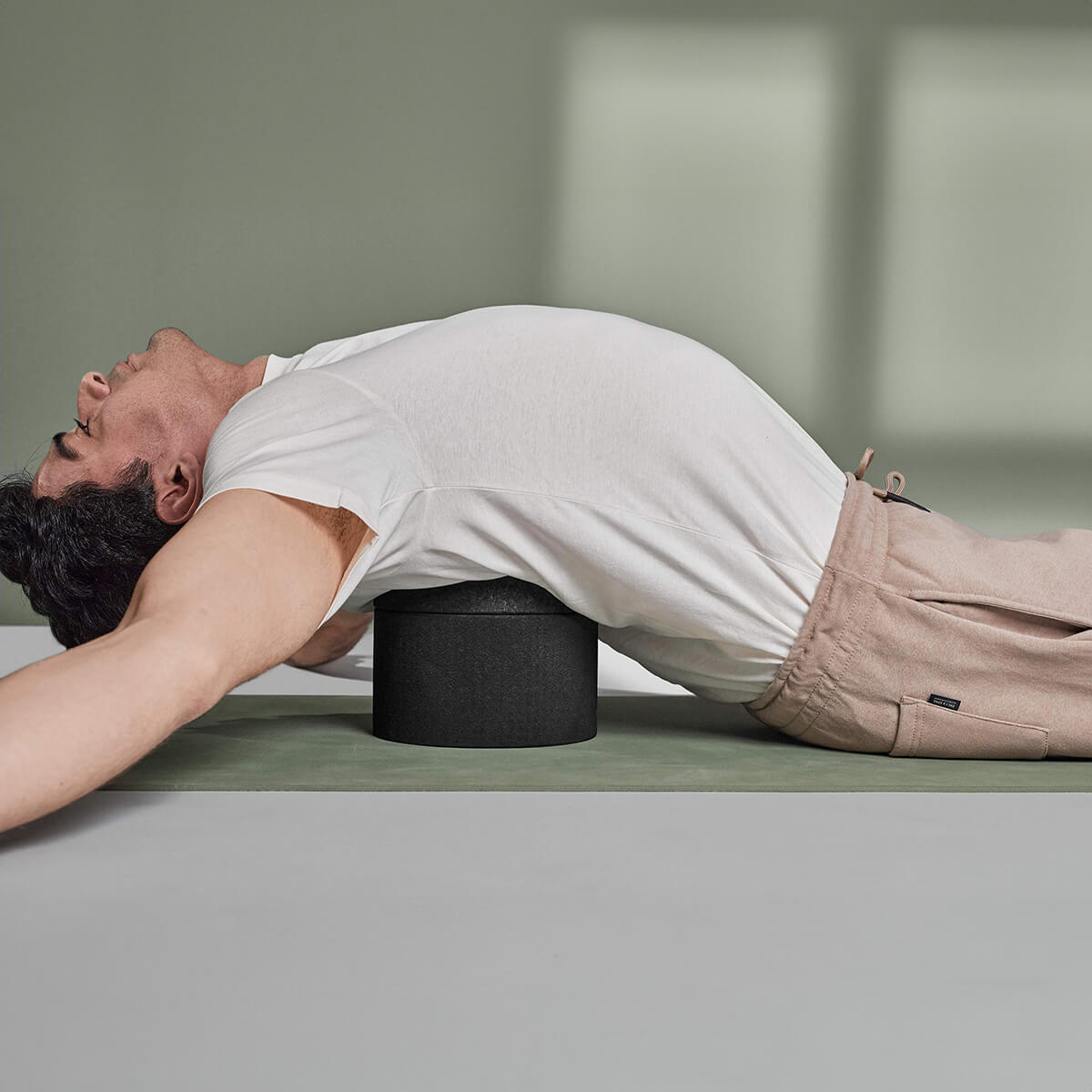 Ein Mann in weißem T-Shirt und beiger Hose liegt auf dem Rücken auf einer Yogamatte, streckt sich mit ausgestreckten Armen und benutzt Der Rückenretter von Liebscher & Bracht unter seinem oberen Rücken zur Mobilisierung und Entspannung der Rückenmuskulatur.