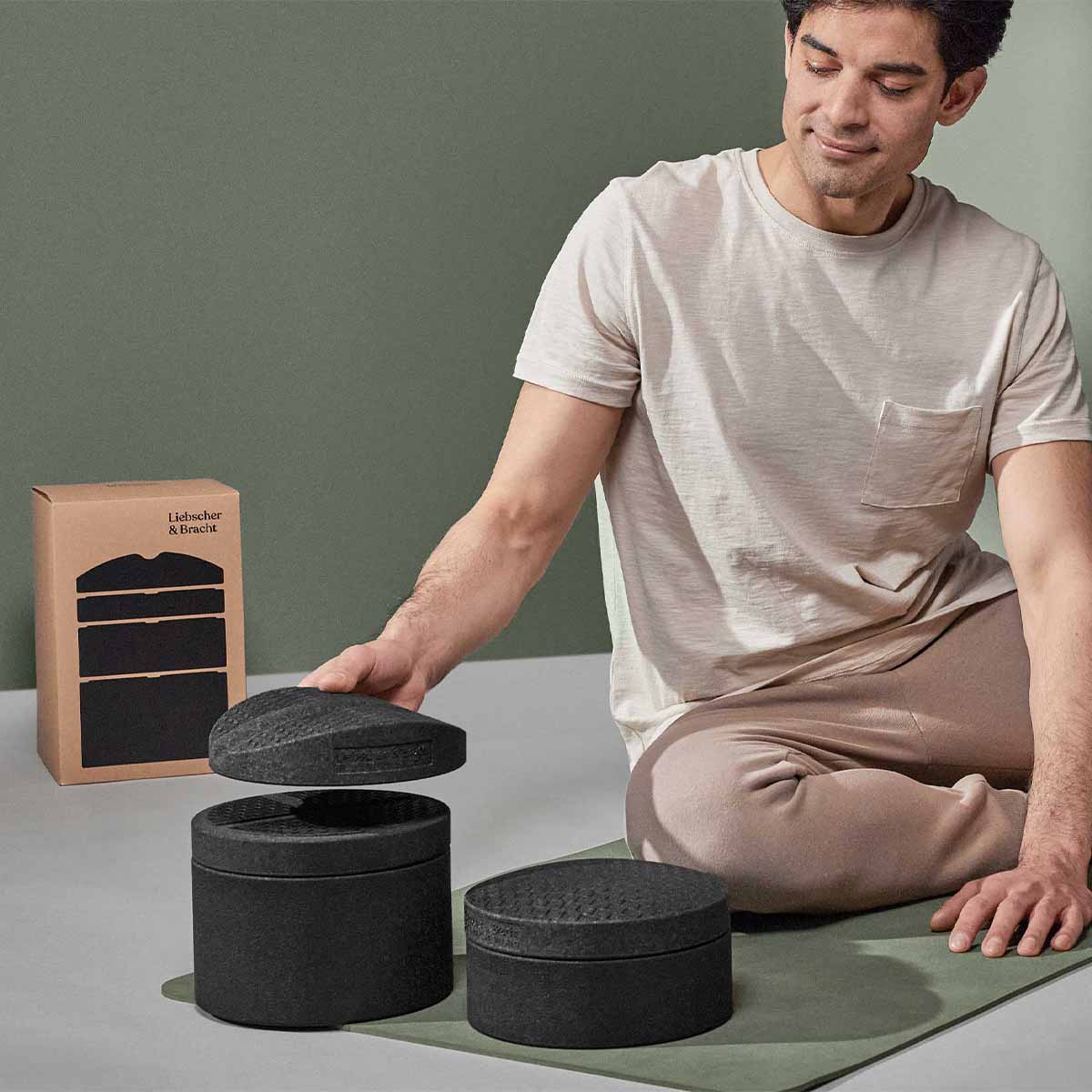 Eine Person in leichter Loungewear sitzt auf einer Yogamatte und stapelt schwarze, strukturierte Schaumstoffblöcke auf oder ab. Im Hintergrund ist eine Markenbox vor einer gedämpften grünen Wand zu sehen.