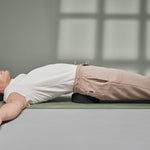 Eine Person liegt auf dem Rücken auf einer Yogamatte, die Arme sind ausgestreckt, sie trägt ein weißes Hemd und eine beigefarbene Hose und benutzt den Liebscher & Bracht Der Rückenretter unter ihrem unteren Rücken zur Mobilisierung und Entspannung der Rückenmuskulatur.