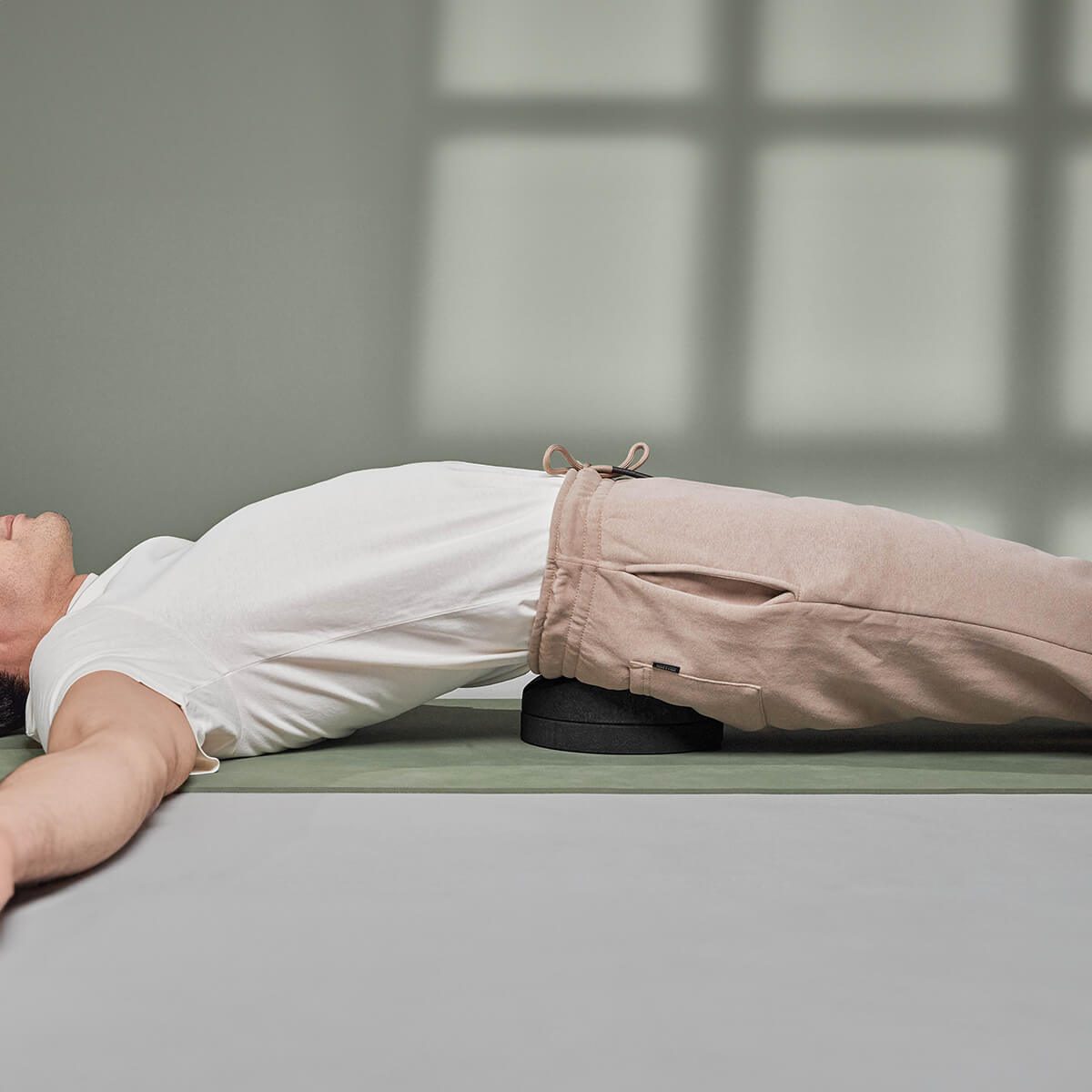 Eine Person liegt auf dem Rücken auf einer Yogamatte, die Arme sind ausgestreckt, sie trägt ein weißes Hemd und eine beigefarbene Hose und benutzt den Liebscher & Bracht Der Rückenretter unter ihrem unteren Rücken zur Mobilisierung und Entspannung der Rückenmuskulatur.