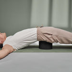 Eine Person liegt auf dem Rücken auf einer Yogamatte, hat die Arme ausgestreckt und benutzt Der Rückenretter von Liebscher & Bracht unter dem unteren Rücken, um die Entspannung und Mobilisierung der Rückenmuskulatur zu unterstützen. Sie trägt ein weißes Hemd und eine beige Hose.