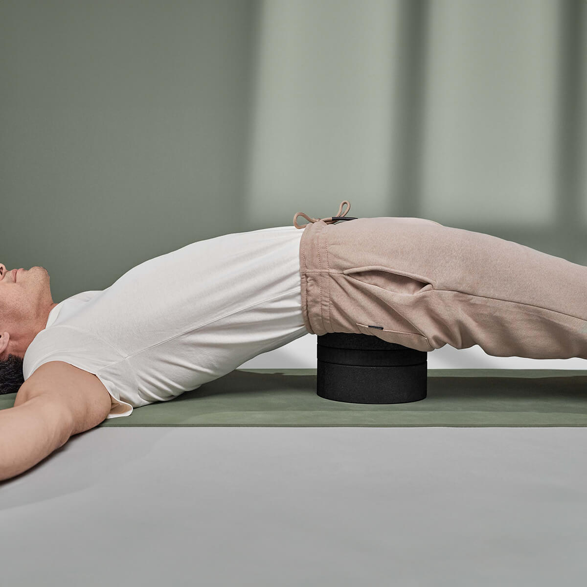 Eine Person liegt auf dem Rücken auf einer Yogamatte, hat die Arme ausgestreckt und benutzt Der Rückenretter von Liebscher & Bracht unter dem unteren Rücken, um die Entspannung und Mobilisierung der Rückenmuskulatur zu unterstützen. Sie trägt ein weißes Hemd und eine beige Hose.