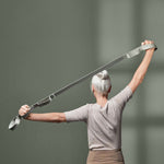 Ein älterer Erwachsener mit grauem Haar in einem Dutt hält ein Spannband über dem Kopf, die Arme ausgestreckt, und steht vor einem schlichten grünlichen Hintergrund. Die Person ist von hinten zu sehen.