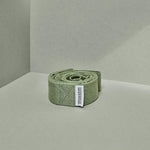 Ein aufgerollter, olivgrüner Stoffgürtel Der Schulterretter von Liebscher & Bracht mit weißem Etikett liegt auf hellgrünem Grund in der Nähe der Kreuzung zweier Wände.