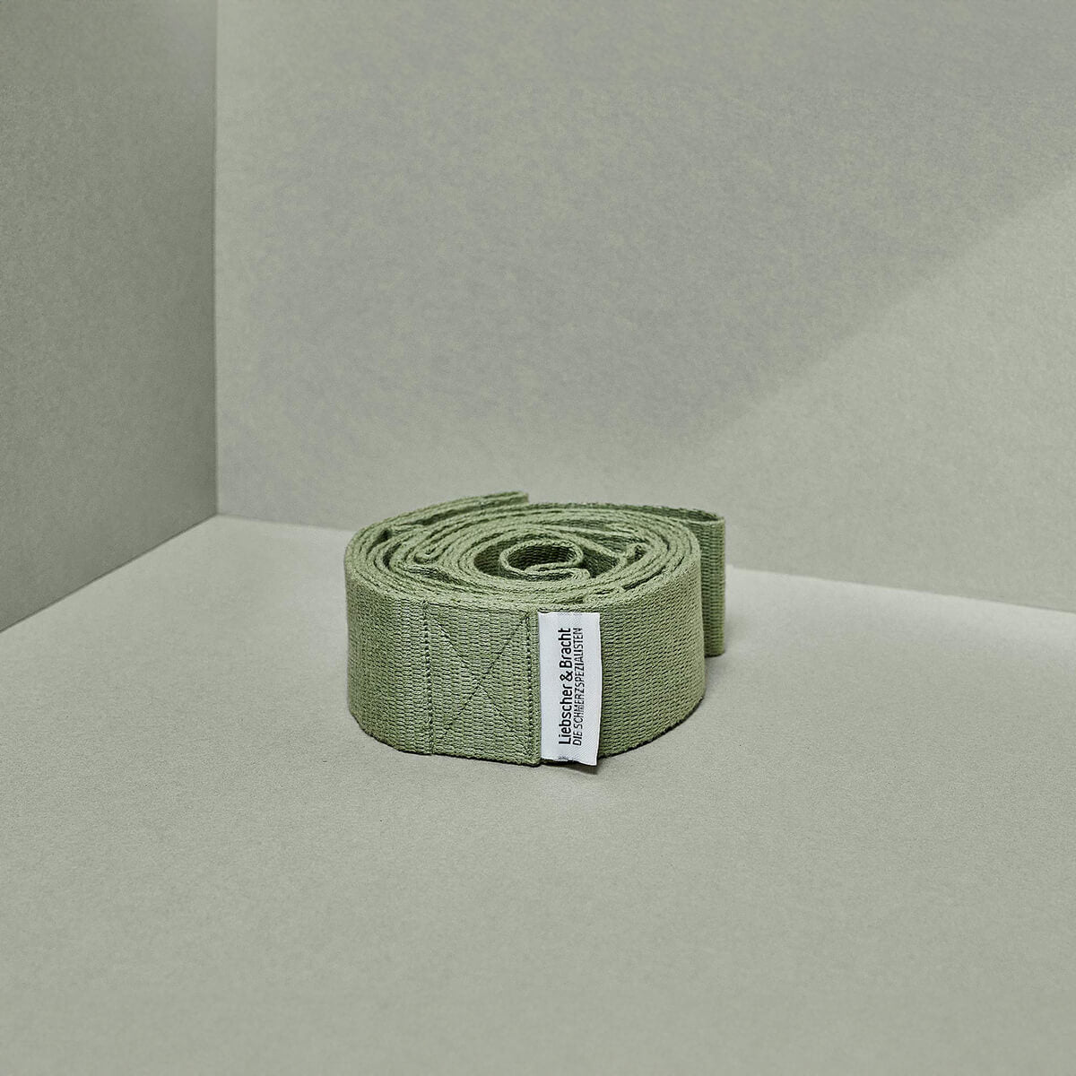 Ein aufgerollter, olivgrüner Stoffgürtel Der Schulterretter von Liebscher & Bracht mit weißem Etikett liegt auf hellgrünem Grund in der Nähe der Kreuzung zweier Wände.