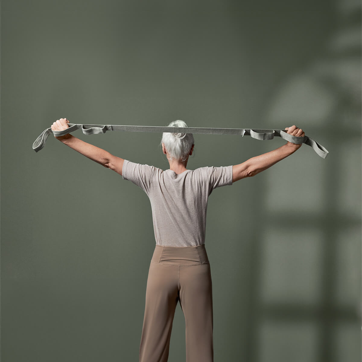 Ein älterer Erwachsener mit kurzen grauen Haaren steht mit erhobenen und ausgestreckten Armen da und hält ein graues Gymnastikband vor einem einfarbig grünen Hintergrund.