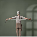 Eine Person mit grauen Haaren nutzt Der Schulterretter von Liebscher & Bracht für das Schultertraining. Sie dehnt ein Widerstandsband gegen eine grüne Wand, während sie ein helles Hemd und beigefarbene Leggings trägt und das Sonnenlicht ihren Schatten wirft.