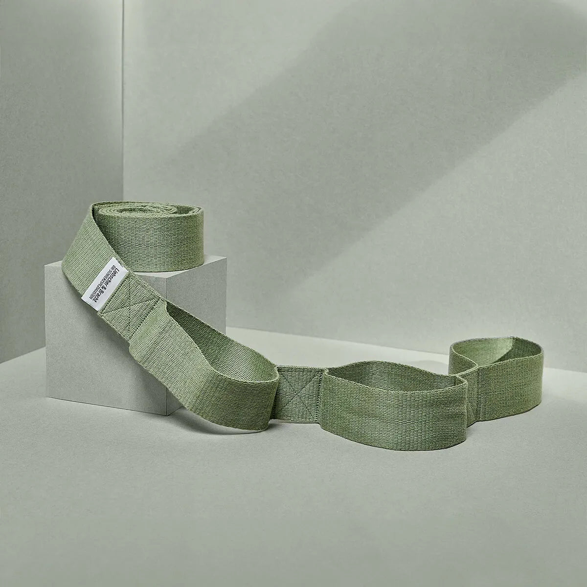 Der Schulterretter von Liebscher & Bracht, ein sagegrünes Gewebeband mit regulierbarer Dehnung, Ziernähten und kleinem weißen Label, ist über einen hellgrauen geometrischen Block vor hellem Hintergrund drapiert.