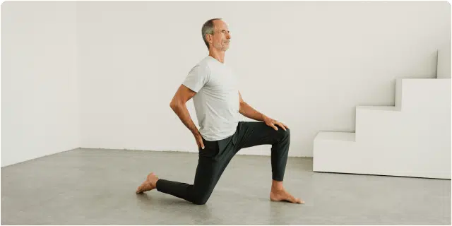 Ein Mann in weißem T-Shirt und schwarzer Hose führt eine kniende Ausfallschrittübung auf einem glatten Boden aus, wobei er ein Knie nach vorne beugt und die Hände auf die Hüften stützt. Im Hintergrund ist eine weiße Treppe zu sehen.