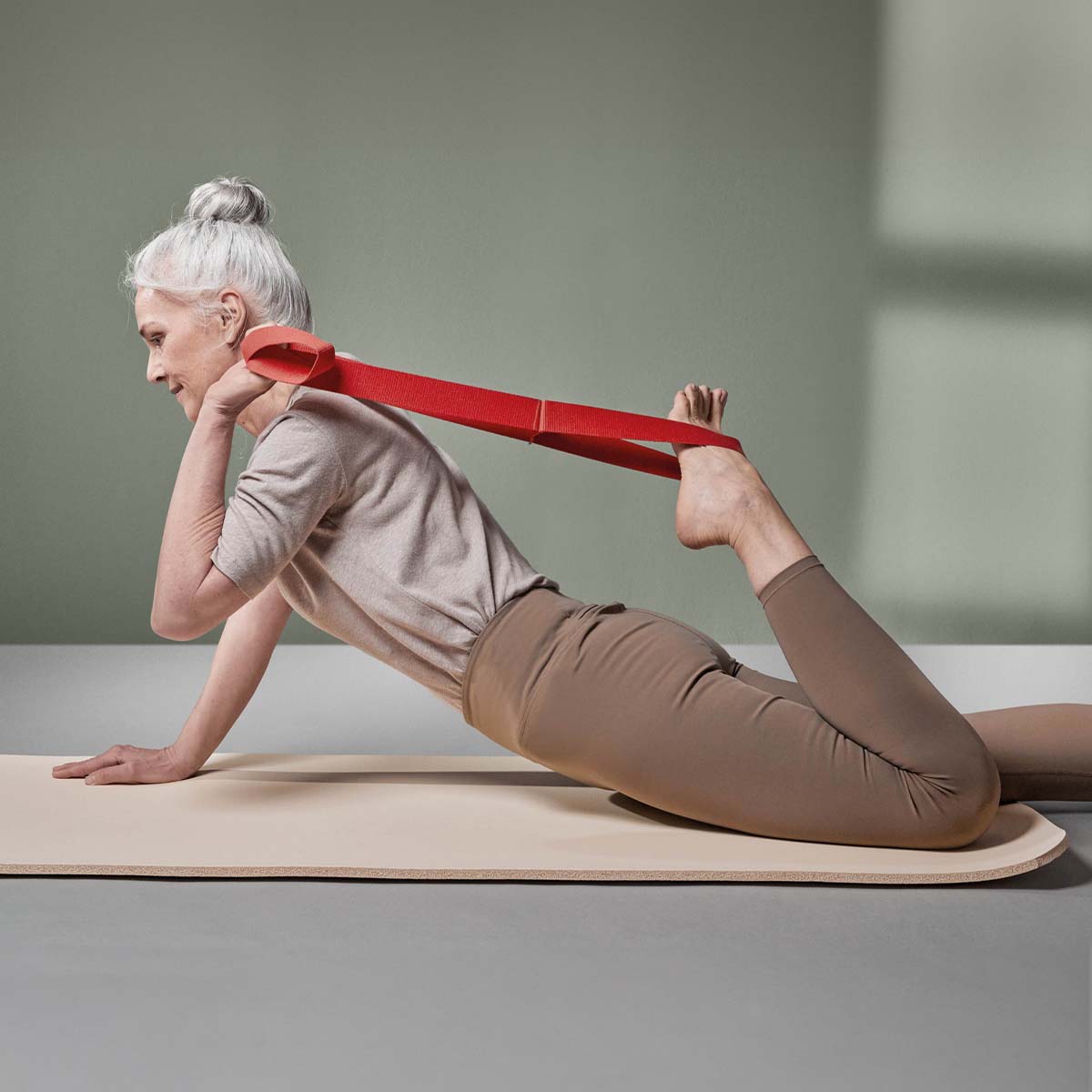 Ältere Frau mit grauem Haar in einem Dutt übt Dehnpositionen auf einer Matte, wobei sie die rote Liebscher & Bracht Übungs-Schlaufe benutzt, um ihren erhobenen Fuß hinter ihrem Rücken zu halten, in einem hellen minimalistischen Raum, um die Beweglichkeit zu verbessern.